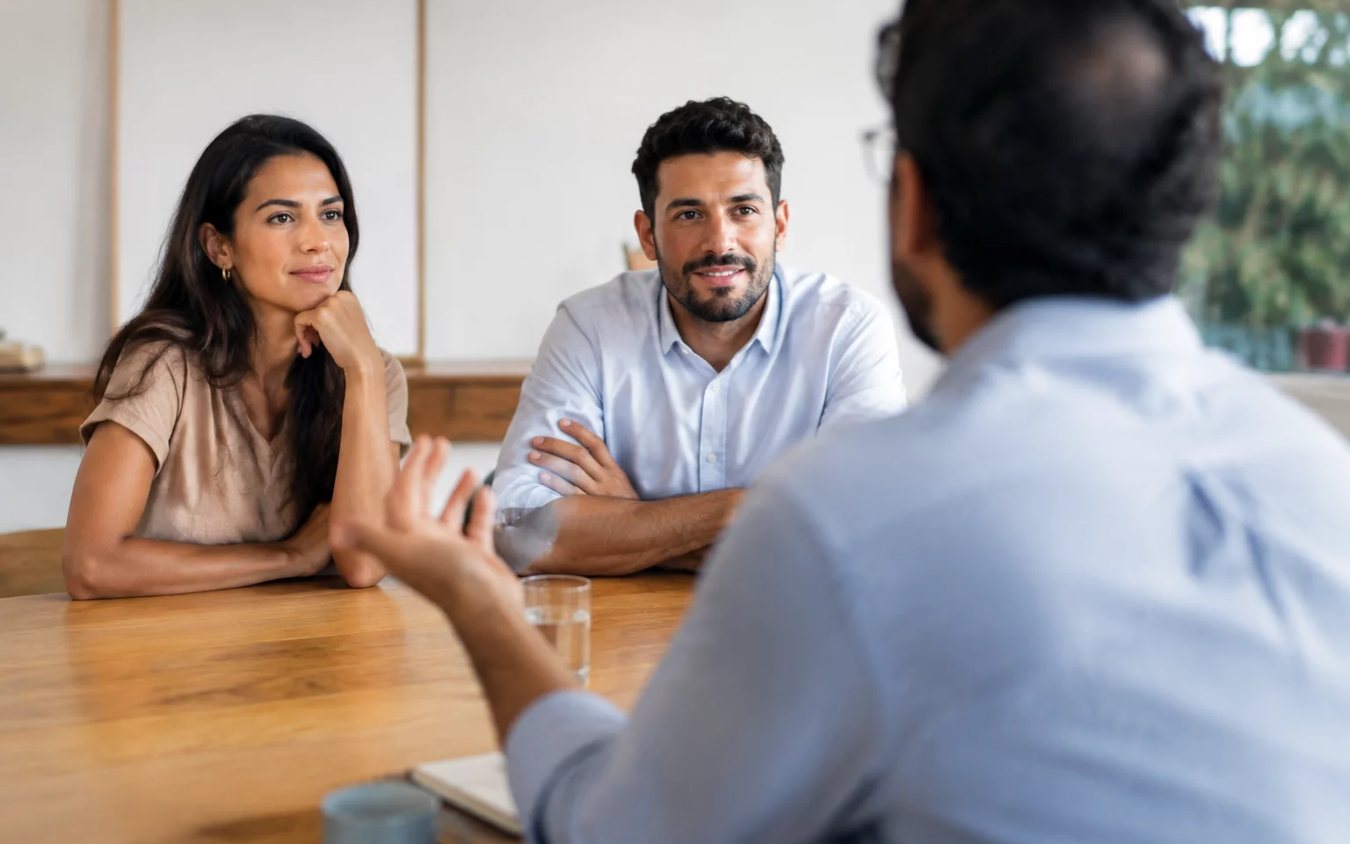 Três participantes conversam ao redor de uma mesa comprida numa sala clara de janelas altas